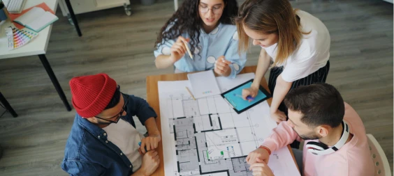Equipo de cuatro profesionales revisando planos arquitectónicos sobre una mesa, mientras colaboran con una tableta y documentos.