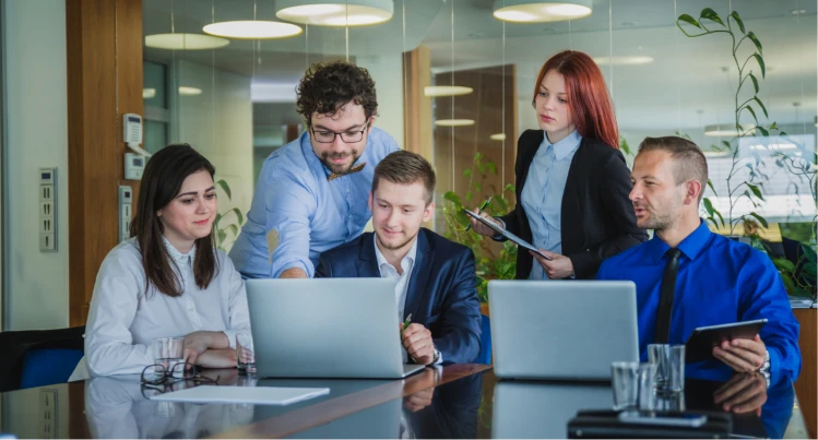 Equipo de cinco profesionales colaborando alrededor de una mesa con computadores portátiles y una tableta en una oficina moderna.