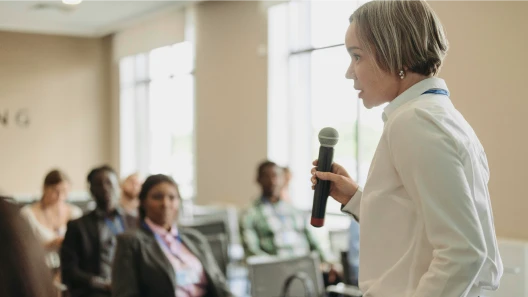 Mujer realizando una presentación con micrófono frente a un grupo de asistentes en una sala de conferencias.